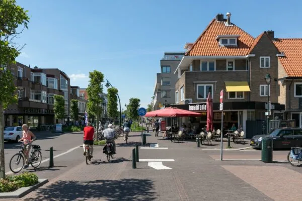 Fietsers en voetgangers bewegen zich door de zonnige winkelstraat 1e Hogeweg met terras en winkels, waaronder Kwalitaria.